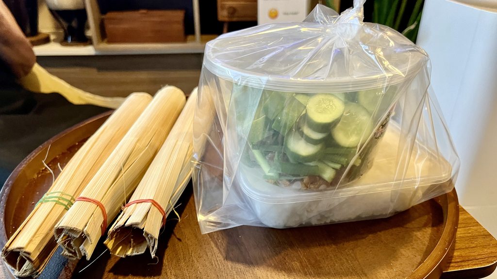 テーブルにカオラムと使い捨て容器に入ったご飯ときゅうりなどのおかず、 ビニール袋に包まれている